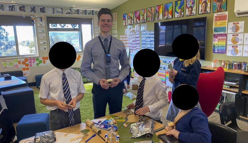 A man in a classroom smiles at the camera, four students beside him (faces obscured)