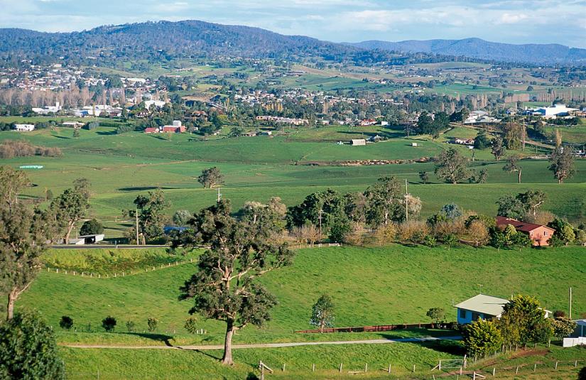 Daytime landscape image of green fields, trees and distant hills. Some houses and buildings scattered throughout.