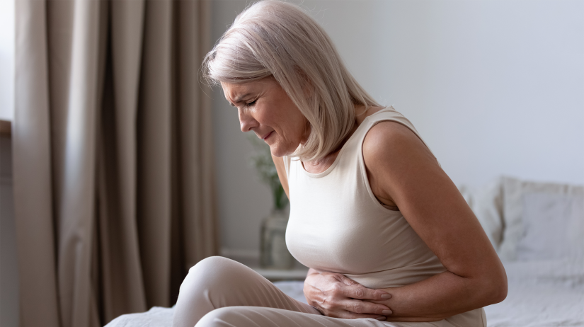 Senior warmer with blonde hair sits on bed clutching abdomen with pained facial expression