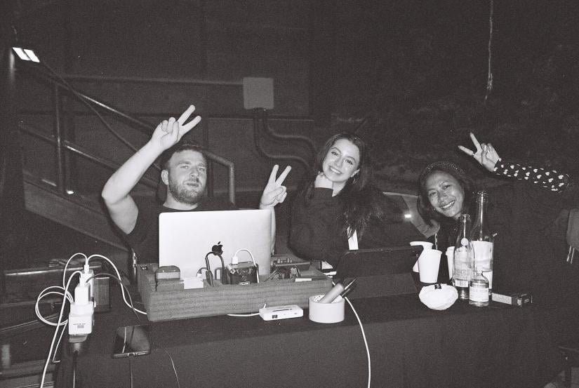black and white image of grace and two other people at a desk with a female and male. They are smiling at the camera and giving 'peace fingers'