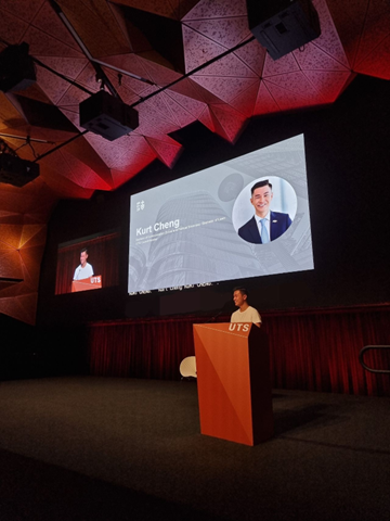 Image of student at lectern giving a lecture standing in front of big screen with image of the speaker