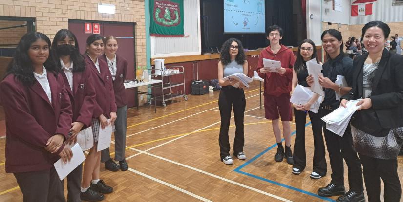 Image taken inside high school gym. University students and academics standing with high school students in maroon uniforms. All are smiling at the camera.