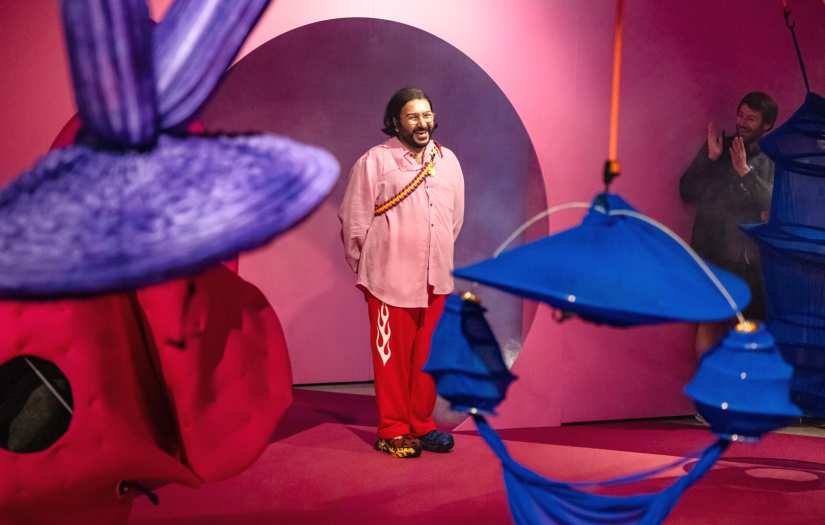 A smiling man, hossein, stands amongst his vibrant art installations as onlookers applaud