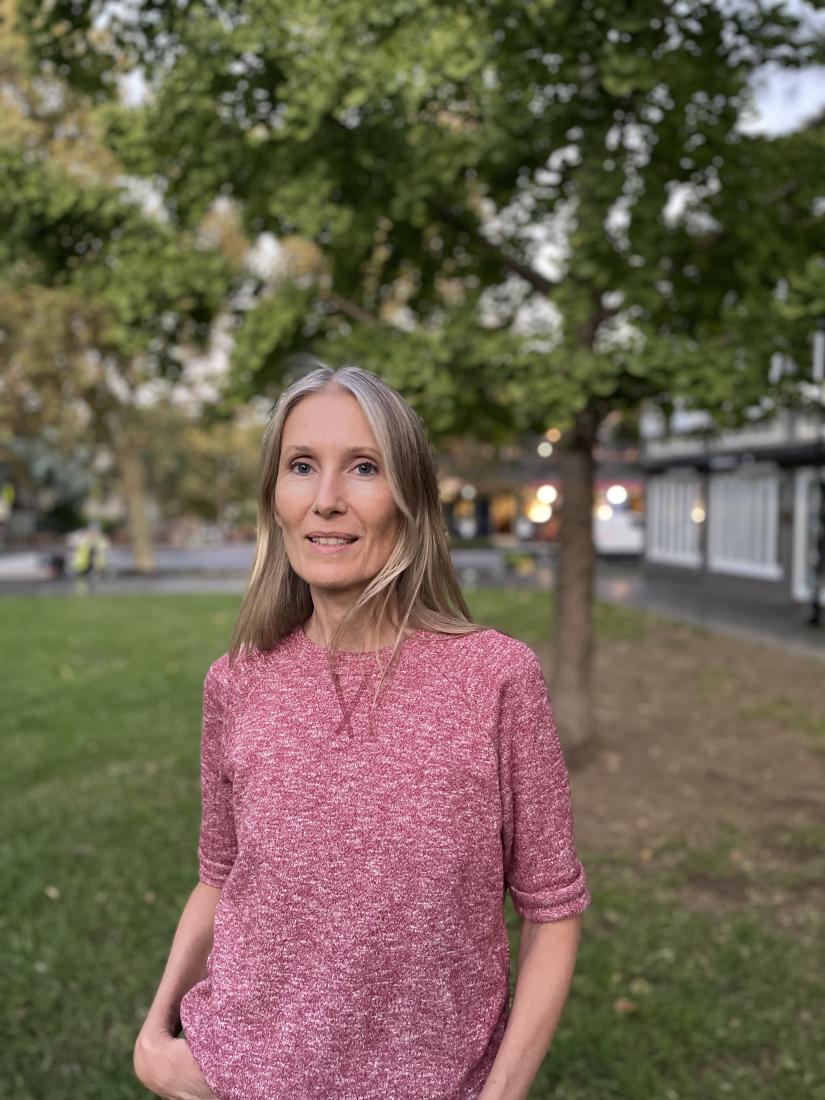Tracy Ellis stands outdoors in front of a tree, wearing a pink shirt and smiling at the camera