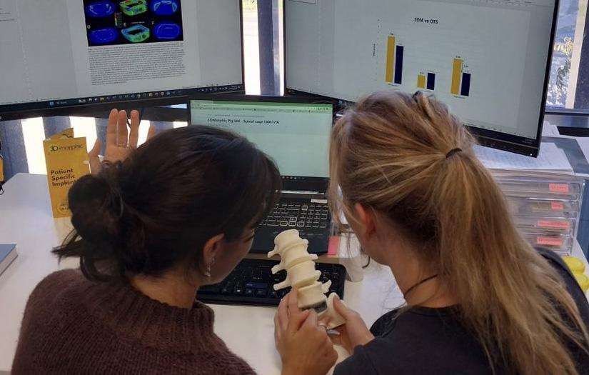 Two girls sitting at a desk in front of a laptop and two screens, and holding a piece of vertebra