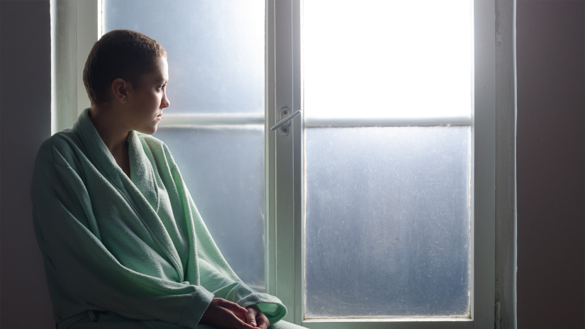 Young woman with short hair sits in front of window. She is wearing a bath robe and looking out the window, which has frosted glass.