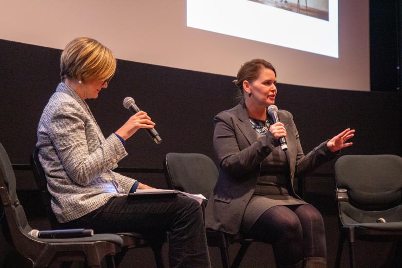 Two women speak on stage, both holding microphones