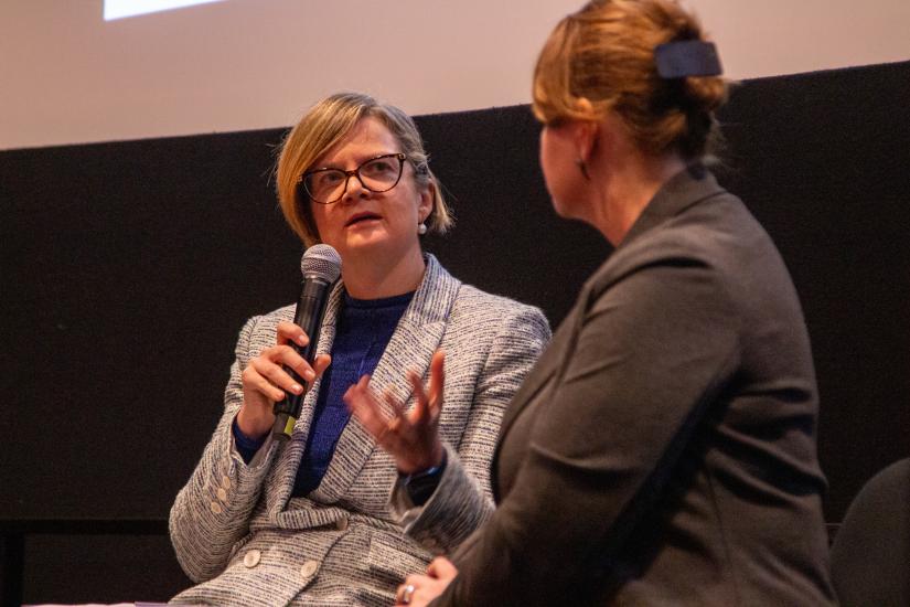 A woman holds a microphone while speaking and gesturing to another woman on stage
