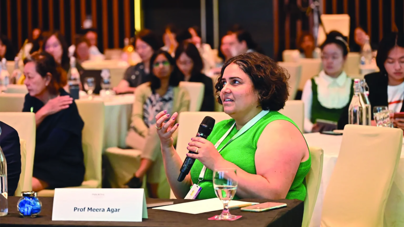 Profesor Meera Agar is sitting at a table and speaking into a microphone. She is wearing a bright greensleevless top and has a lanyard around her neck. Her name is on a card on the table in front of her. Other delegates are blurred in the background.