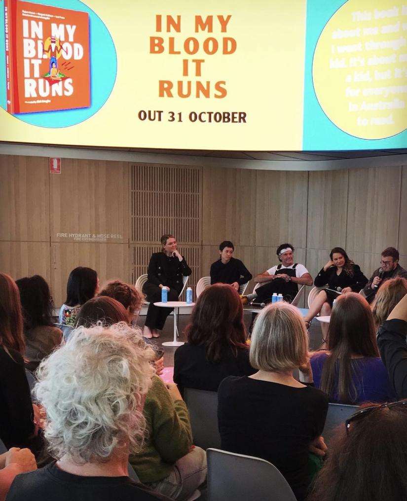 A panel sits discussing underneath a sign promoting In My Blood It Runs