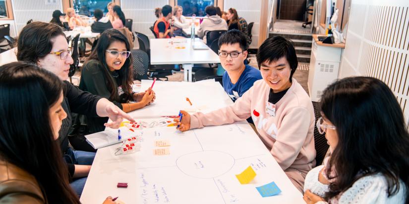 Six students in the midst of a discussion sit around a table in a brightly lit classroom. On the table is a large sheet of paper with sticky notes and writing.
