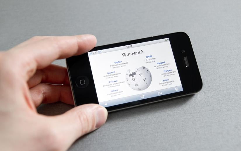 A person holds a mobile phone which displays the Wikipedia homepage.
