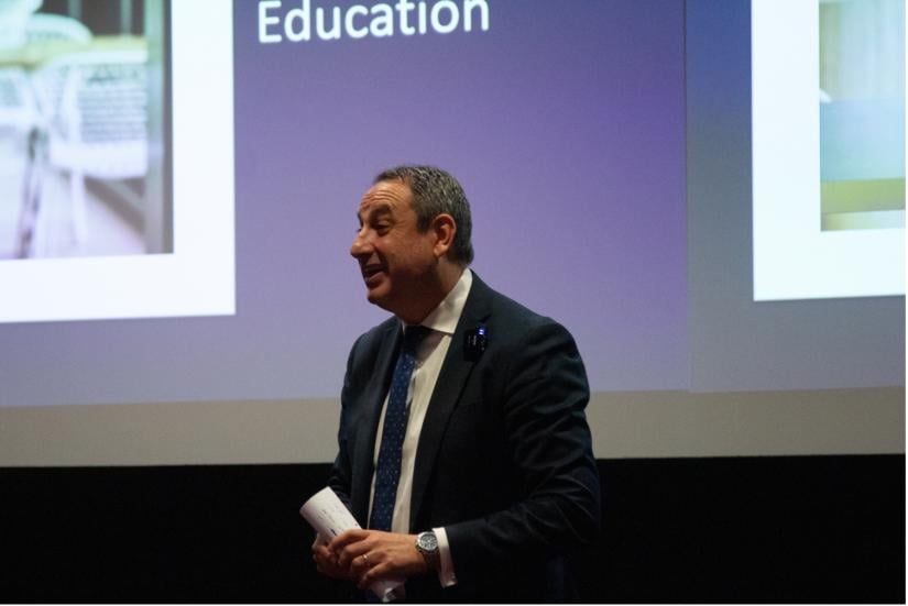 A man in a suit speaks to a crowd, smiling