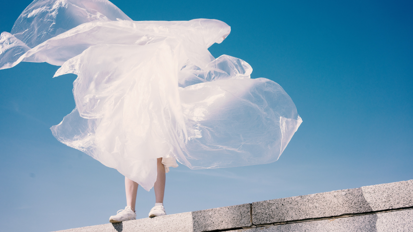 Feet in sneakers standing on a wall in front of blue sky, person appears to be wearing a large piece of plastic that is floating in the breeze.