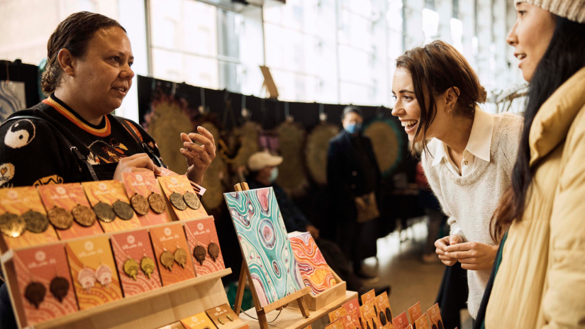 Two women interact with a stallholder at an art fair.