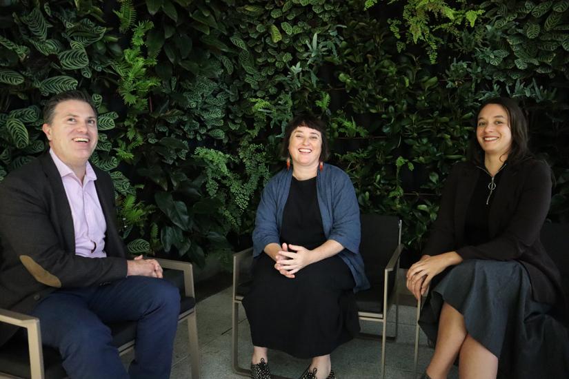 Three people sitting in front of a green plant background. On the left a man is smiling and looking at the camera wearing a business shirt and jacket. On the right of the image are two UTS researchers sitting next to each other. The one in the centre of the image is a woman with short dark hair wearing a black dress and denim jacket. The one on theright of the image has shoulder length dark hair and is wearing a black top and skirt.