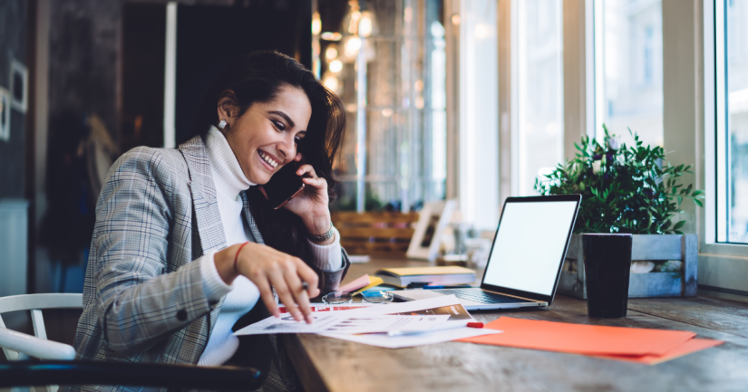 A women in a grey jacket smiles as she talks on the phone and looks down at paper reports and her laptop.