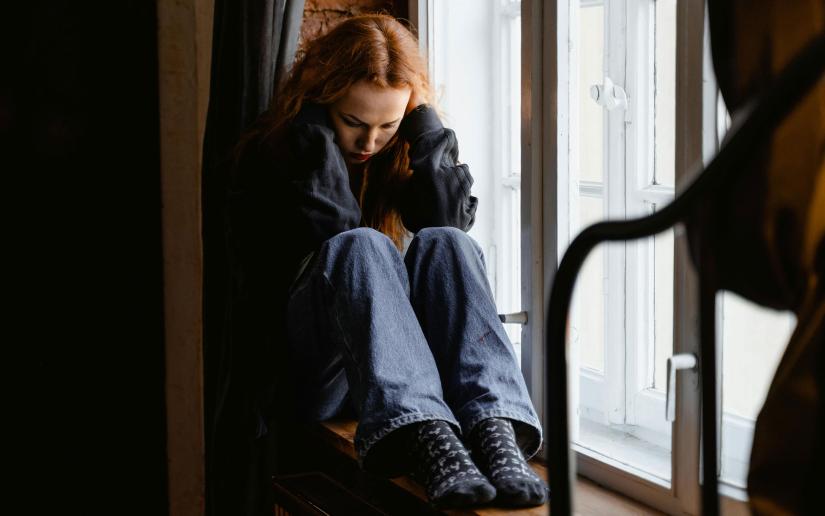 Stock picture of a young woman sitting by a window with her head in her hands. Mart Production/Pexels