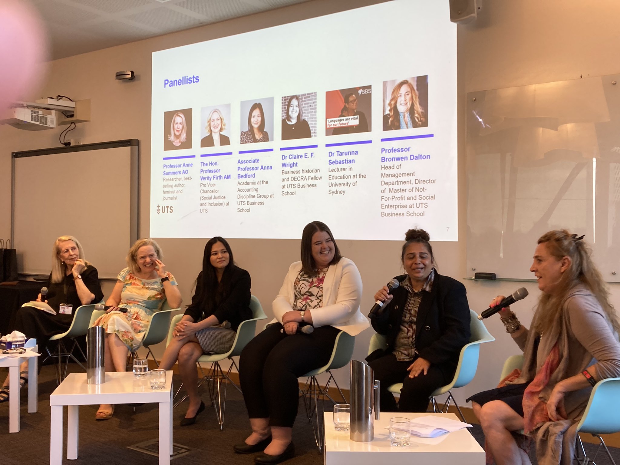The interior of the UTS Business School classroom, at the front of the class is a seated panel seating left to right consisting of Anne Summers, Verity Firth, Anna Bedford, Claire Wright Tarunna Sebastian as Bronwen Dalton moderates with a microphone. Above them a screen displays the names and titles of the panellists. The panel is smiling and laughing as they look to Bronwen who is speaking into the microphone and addressing the unseen crowd.