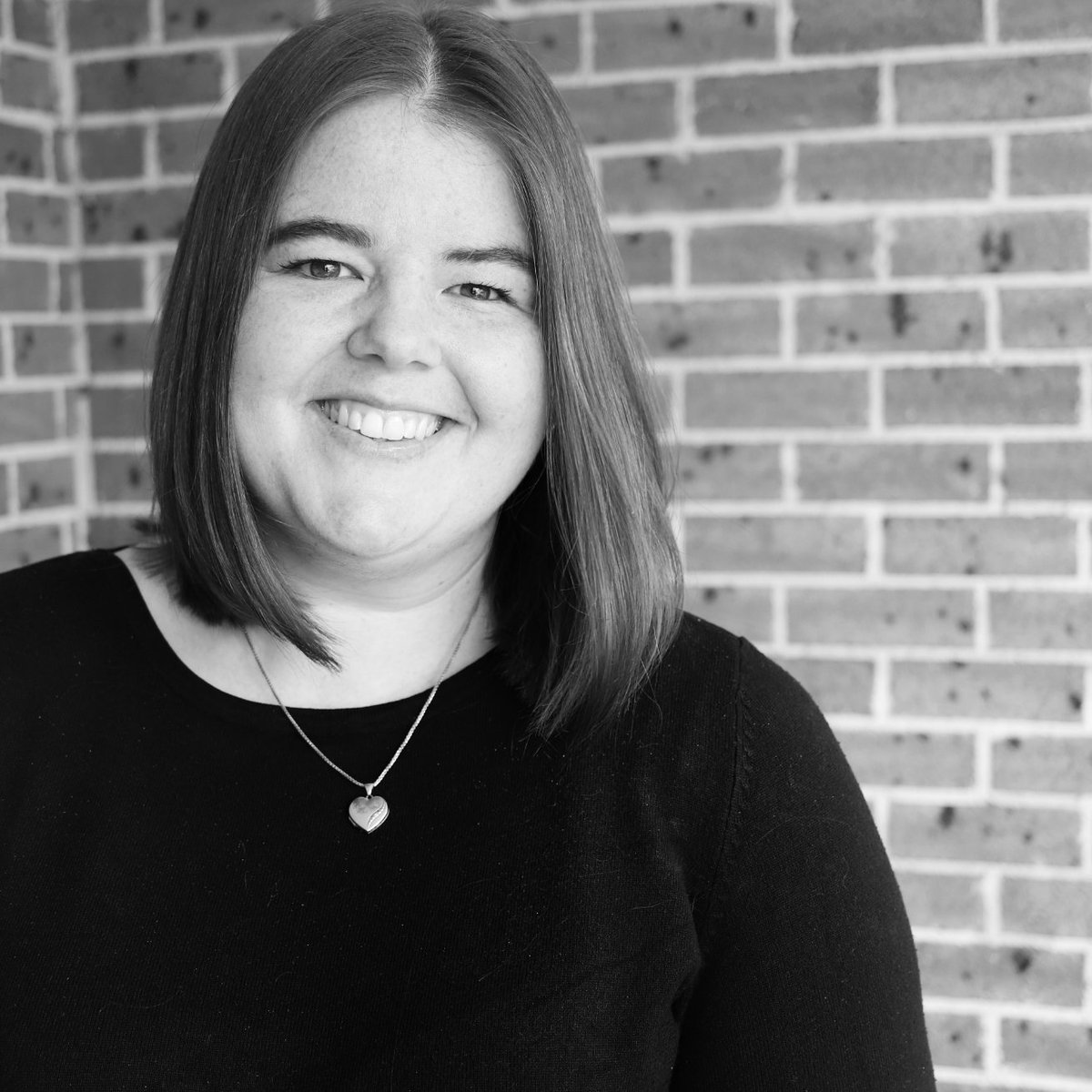Black and white portrait of Claire Wright, she smiles to camera wearing a black top. She wears a silver loveheart shaped necklace. The background is a brick wall.