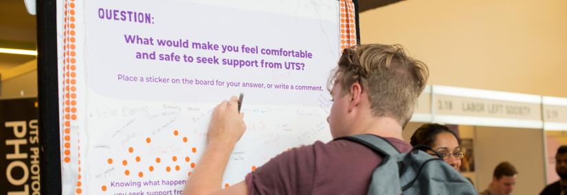 A man in red shirt and blonde hair writes on a board with the question 'What would make you feel comfortable and safe to seek support from UTS?'