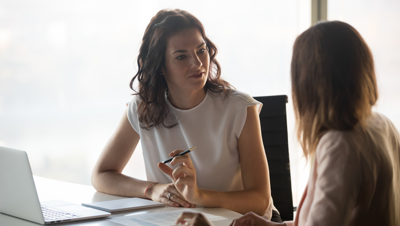 Two women working together at a desk with a laptop. One is facing away from the camera and has shoulder-length brown hair and wears a jacker. The other lady is facing her colleague and has long brown curly hair. She is wearing a short sleeved white top and is holding a pen.