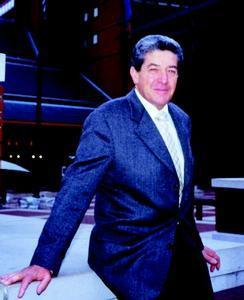 Man (Denis Lenard) in suit and tie seated on the edge of a stage, with a dark auditorium background