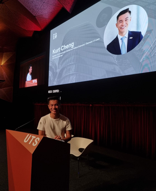 Image of student at lectern smiling at camera. He is standing in front of big screen with image of the speaker