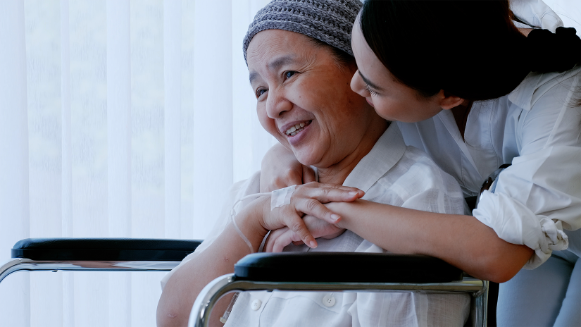 Asian lady with head scarf is sitting in a wheelchair. Her daughter is hugging her from behind.