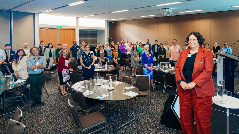 Professor Meera Agar is standing on a stage with the forum delegates in the background