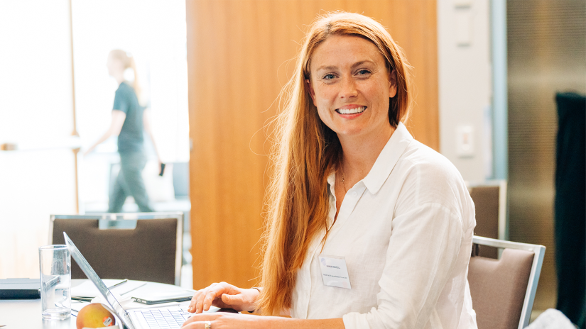 Hannah Wardill is sitting at a conference table with a laptop. She has long red hair and she is wearing a white shirt.