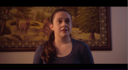 A young woman looking off camera. There is a painting behind her. She has long brown hair.