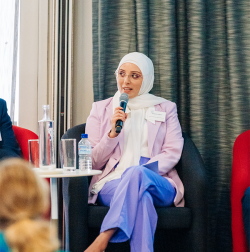 Nadine El-Kabbout is talking with a microphone on a stage at the PaCCSC & CST Annual Research Forum 2023. She is wearing mauve trousers, white shires and pale pink blazer and a white head scarf.