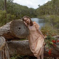 colour image of female in peach dress leaning against fallen log in a forest river setting