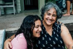 A joyful moment between two women sitting on steps. Both are smiling warmly, embracing each other. The setting appears to be a porch, conveying warmth and contentment.