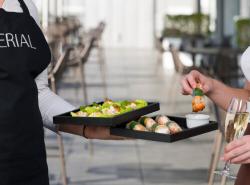 A server carrying trays of small bites while a guest picks up some food and holds a glass of sparkling wine at a catered event.