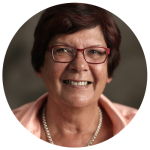 Pat Turner AM smiling. She is wearing a pair of red-framed glasses, a light pink shirt, and a pearl necklace.