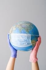 Image of a globe with a disposable mask on it being held between two hands one with a pink glove and one with a blue glove.