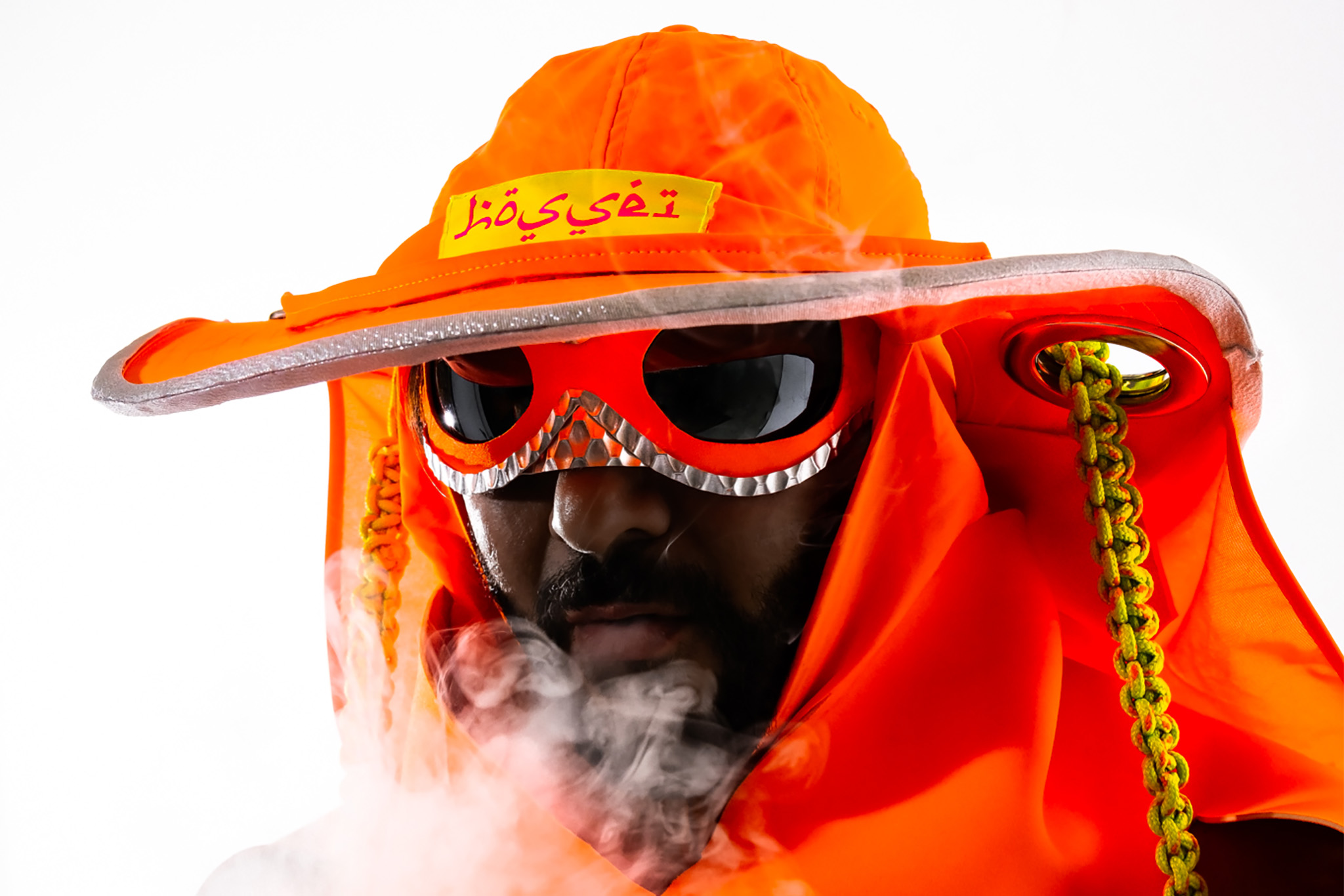 A masked figure wears a bright orange hat and glasses, they blow smoke towards the camera. 