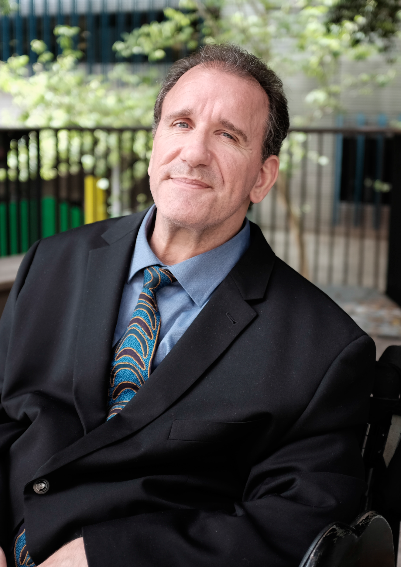 Simon Darcy, sitting, smiling, wearing a light blue button up shirt, a blue tie with brown shapes on it and a black blazer over the top.