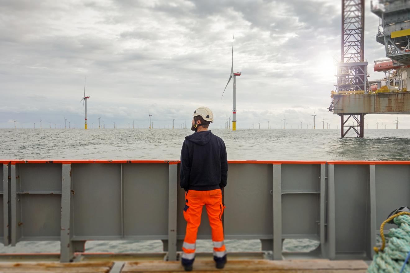 A man stands with his back to us looking out over the sea and a wind turbine