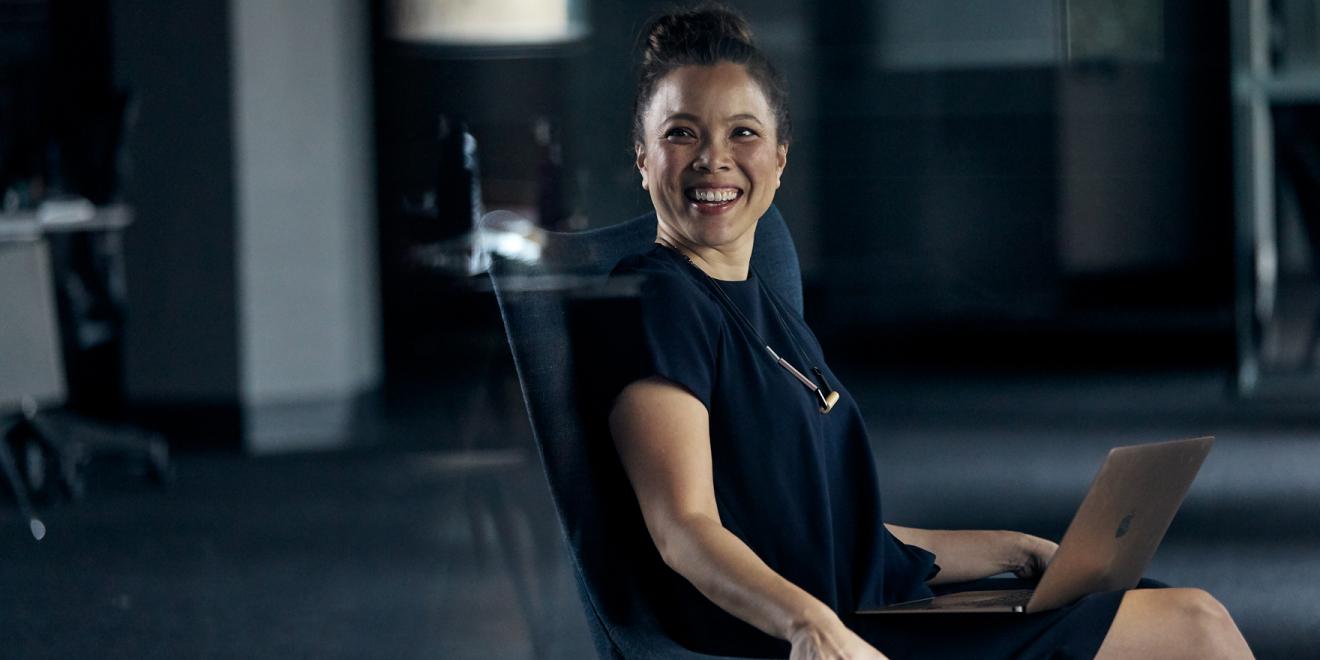 Woman sitting in a chair with hair in a bun wearing a blue business dress and has a silver laptop on her lap