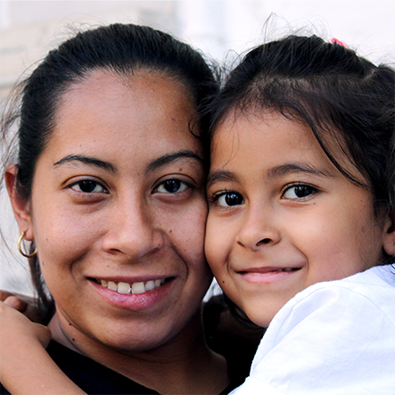 Young daughter embracing mother both with dark hair and eyes looking at camera and smiling