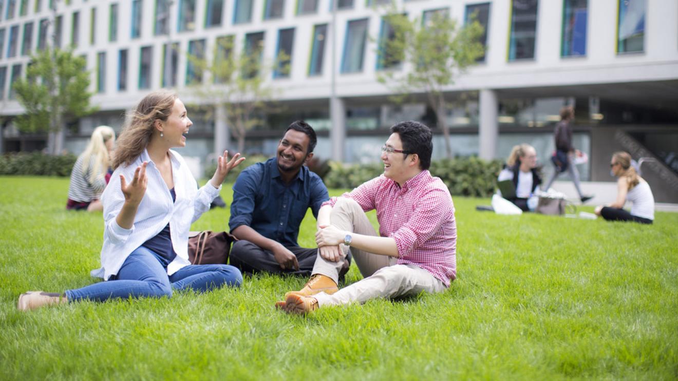Three students sitting and chatting on Alumni Green with other student groups in the background