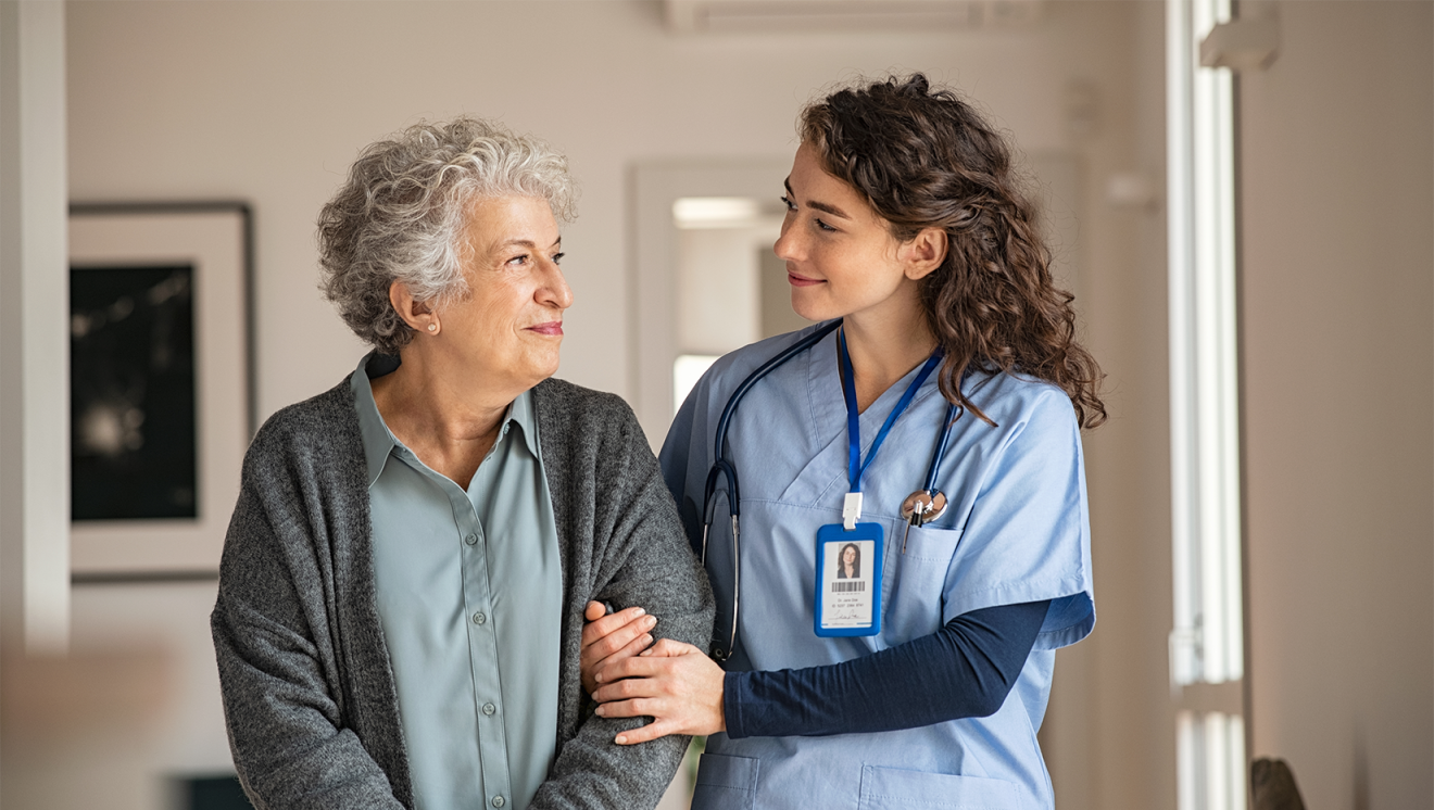 Young nurse with long, brown, curly hair is wearing blue scrubs and walking arm in arm with an older female patient with grey hair who is wearing a sage-green button down shirt and a dark grey cardigan.