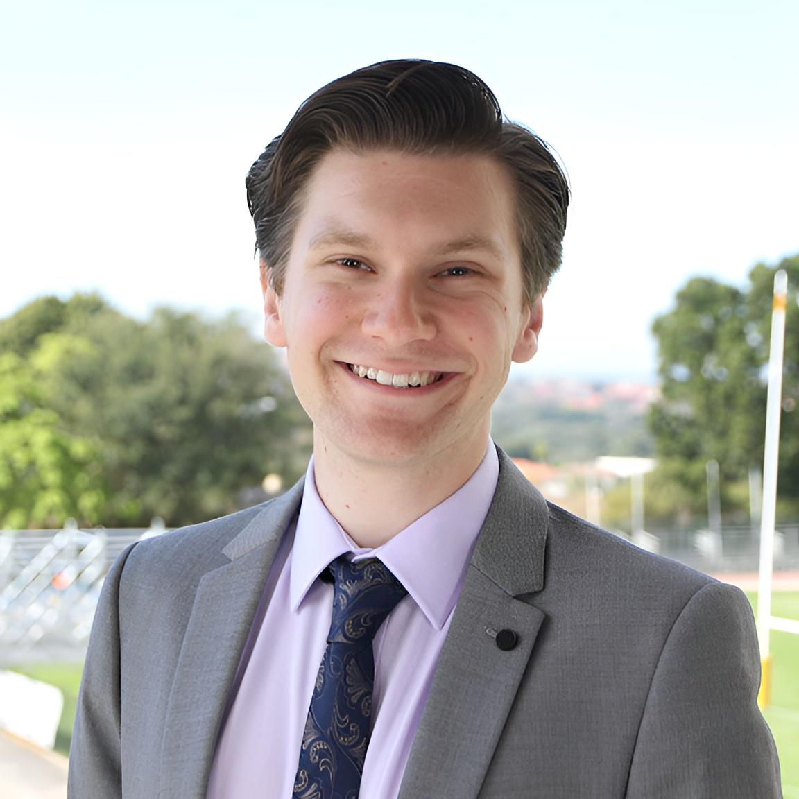 James, wearing a suit and tie, smiles at the camera in front a sports field