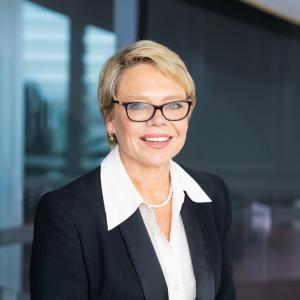 A picture of Sally Cripps, female, short blonde hair, wearing a black glasses, and a suit jacket paired with a white blouse