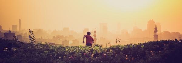 A boy runs away from the camera in a field. A city is just visible in the distance through golden haze.