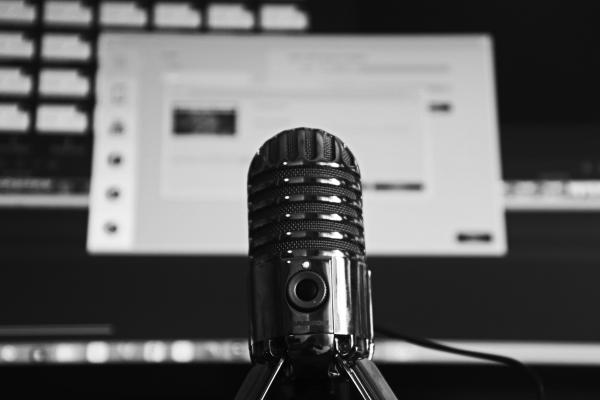Black and white image with a microphone in the foreground and a desktop computer screen in the background.