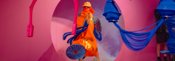 A person wearing a vibrant orange and blue costume surrounded by colourful installations